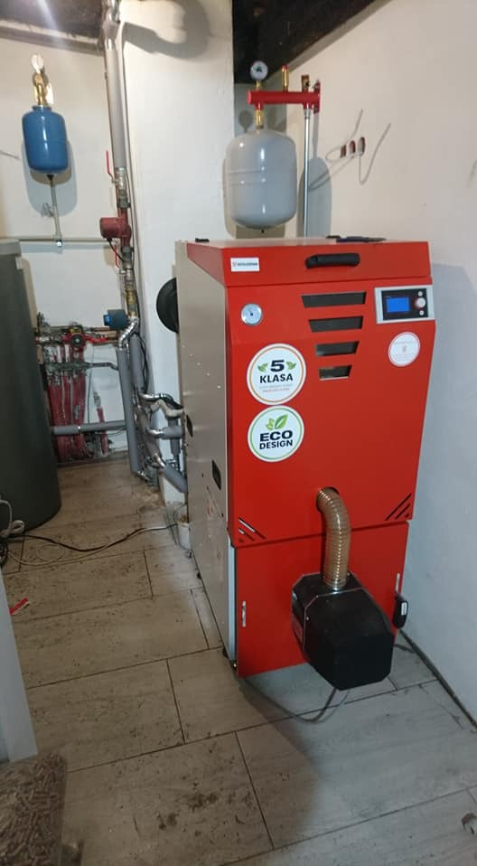 Czerwony piec na paliwo stałe z naklejkami '5 Klasa' i 'Eco Design' w kotłowni z szarą instalacją i zbiornikami wyrównawczymi.