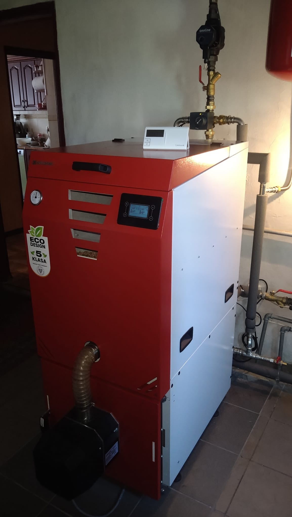 Czerwono-biały piec na paliwo stałe klasy 5 z oznaczeniem Eco Design, z podłączonymi rurami instalacji grzewczej, widoczny w kotłowni.