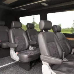 Wnętrze luksusowego busa z czarnymi skórzanymi fotelami, czerwonymi przeszyciami i widokiem na park przez okno. Detal wykończenia premium.