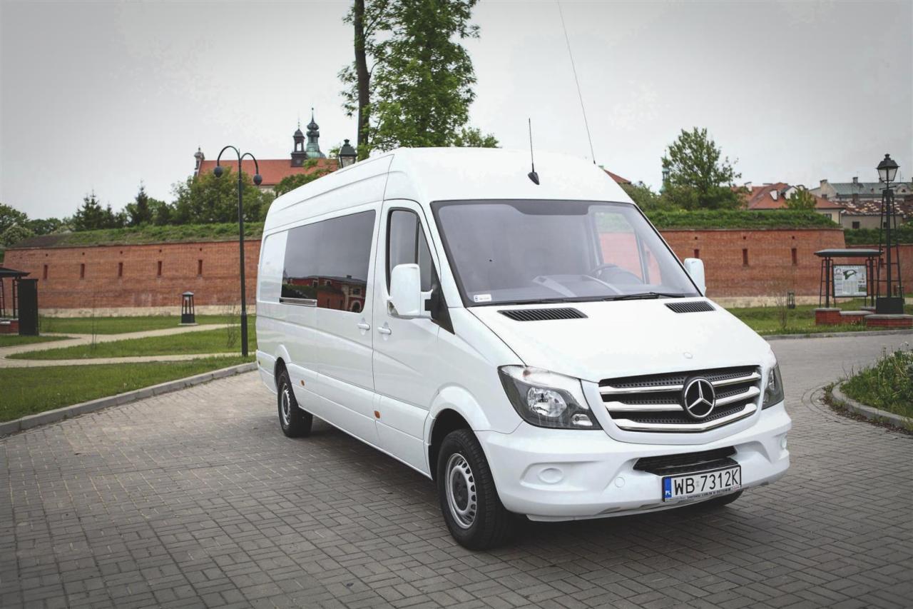Biały bus Mercedes-Benz Sprinter z widoczną tablicą rejestracyjną WB 7312K, zaparkowany na brukowanym placu, z budynkiem w tle. Transport osób.