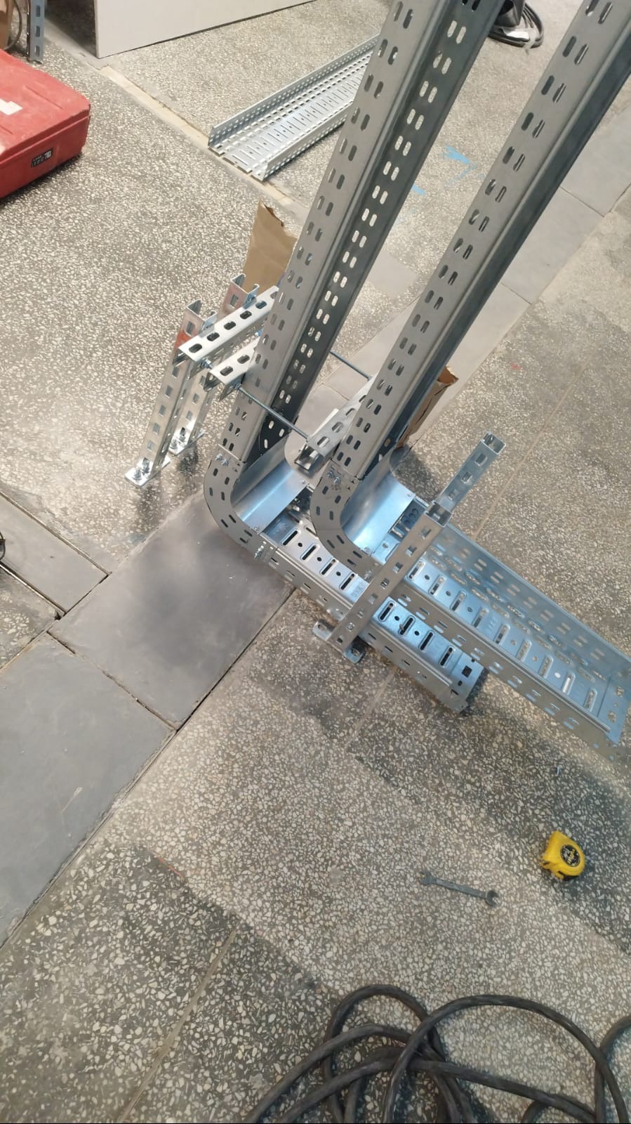 Metalowe korytka kablowe, łączone pod kątem, leżą na podłodze. Widoczny klucz i miarka. Elementy instalacji elektrycznej w trakcie montażu.