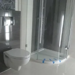 Nowoczesna łazienka z kabiną prysznicową i podwieszaną toaletą. Szare płytki imitujące drewno, białe płytki i podłoga. Widoczne taśmy zabezpieczające kabinę.