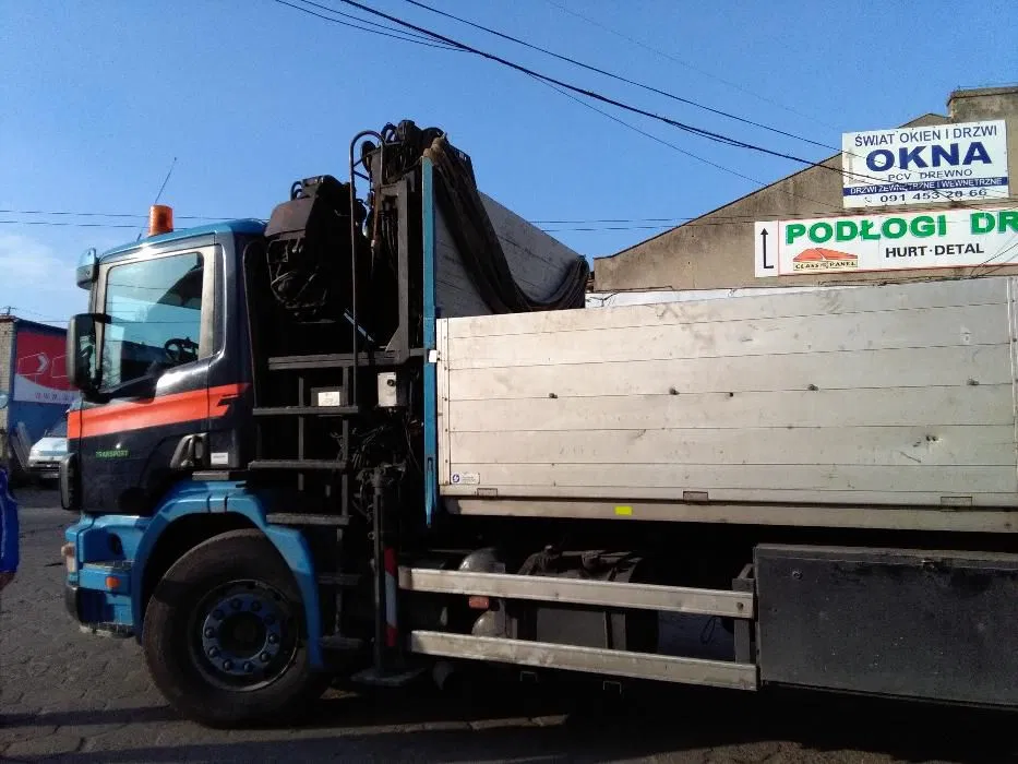 Ciężarówka z dźwigiem hydraulicznym na tle budynku z reklamami okien i podłóg, widok z boku, niebiesko-czarna kabina z pomarańczowym pasem.