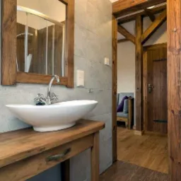 Rustykalna łazienka z drewnianą umywalką, lustrem w drewnianej ramie i szarą glazurą. Widoczne drewniane elementy konstrukcyjne i drzwi. Styl retro.
