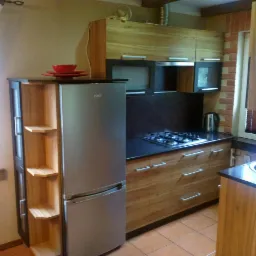 Nowoczesna kuchnia z drewnianymi frontami, czarnymi blatami i srebrną lodówką z półkami bocznymi. Widoczna płyta gazowa i okno z drewnianymi żaluzjami.