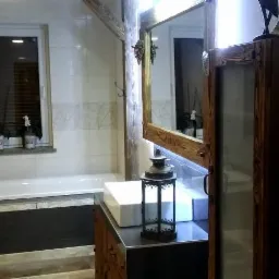 Rustykalna łazienka z drewnianymi elementami, białą armaturą i dekoracyjną latarnią. Widoczne lustro w drewnianej ramie i okno z roślinami.