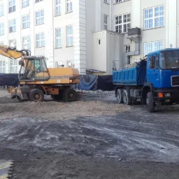 Żółta koparka kołowa MAN 1183 i niebieska wywrotka MAN na placu budowy przed budynkiem z licznymi oknami.