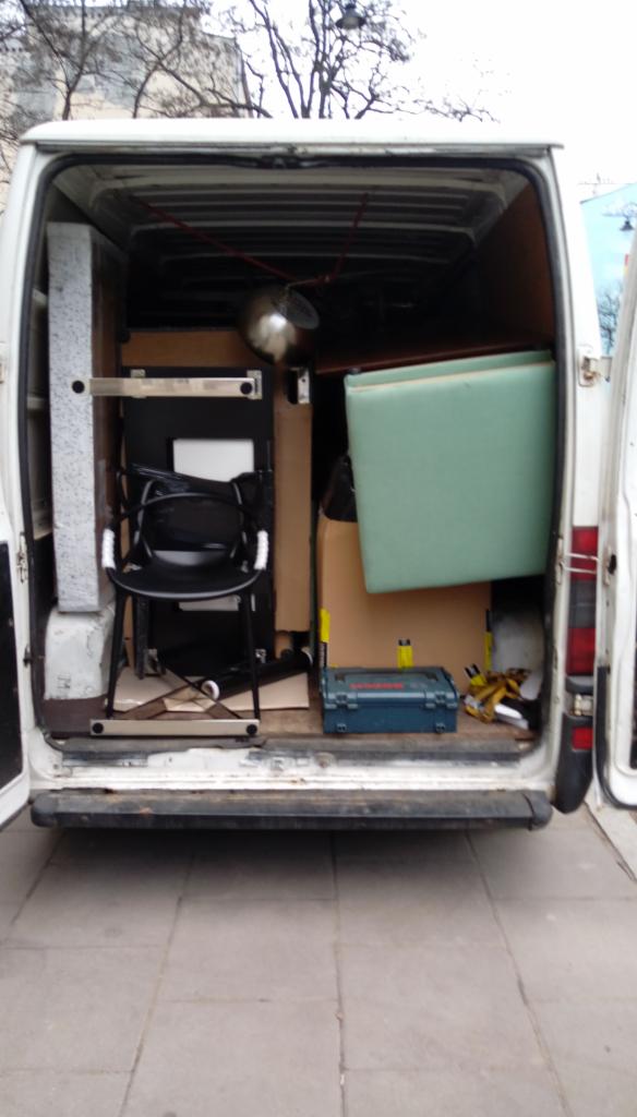 Wnętrze białego busa załadowane meblami, kartonami i narzędziami, widok z tyłu, otwarte drzwi.