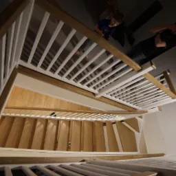 Widok z góry na drewniane schody z białymi balustradami i drewnianymi poręczami. Na górze schodów widoczne dwie osoby.