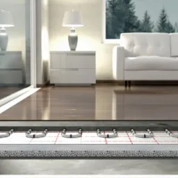 Przekrój warstw podłogi z ogrzewaniem podłogowym w Legnicy, widoczne rury grzewcze zamocowane na macie z siatką, izolacja oraz wylewka, w tle salon z drewnianą podłogą.