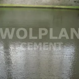 Świeżo wylana, mokra posadzka cementowa marki Wolplan, z widoczną taśmą dylatacyjną przy ścianie i odbiciami światła na powierzchni.