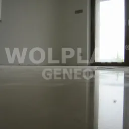 Błyszcząca, gładka posadzka w jasnym wnętrzu w Legnicy. Widoczne okno balkonowe i fragment ściany. Podłoga odbija światło, tworząc lustrzany efekt.