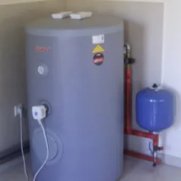 Szary bojler Galmet z podłączonym osprzętem (rury, manometr, zabezpieczenia) stoi w rogu pomieszczenia na jasnej podłodze. Widoczne gniazdko elektryczne.