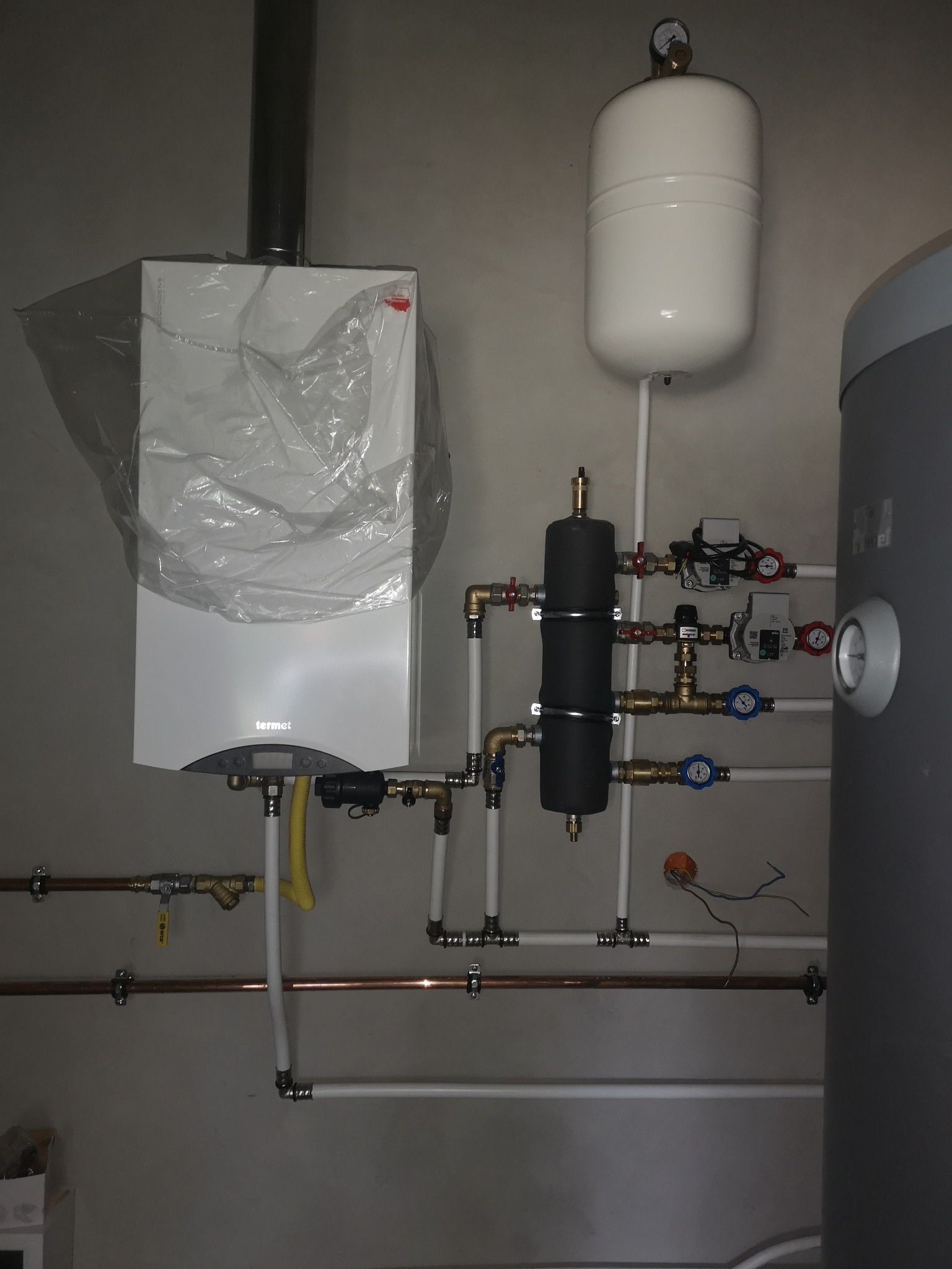 Nowy piec gazowy marki Termet, jeszcze w folii ochronnej, podłączony do instalacji centralnego ogrzewania z zbiornikiem wyrównawczym i zasobnikiem CWU, widoczne liczne rury miedziane i białe PCV...