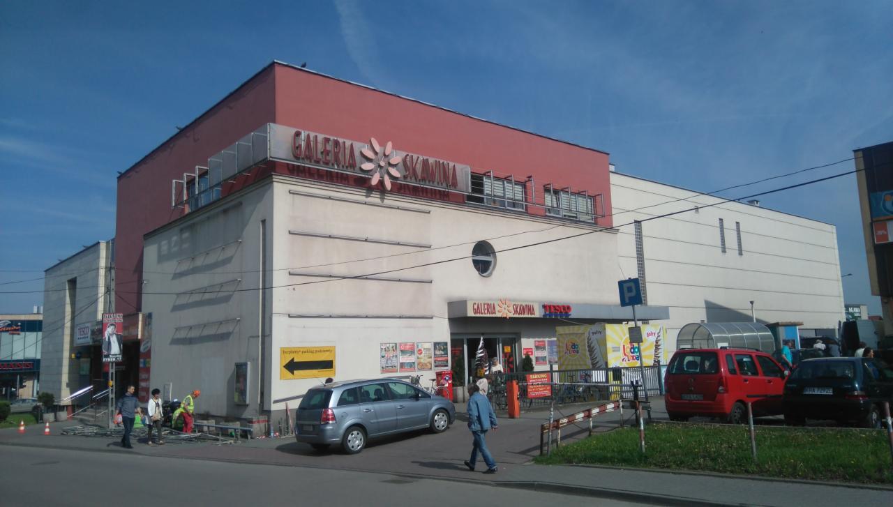 Widok na budynek Galerii Skawina z elewacją w odcieniach bieli i czerwieni, z widocznym logo i wejściem, na tle błękitnego nieba, z zaparkowanymi samochodami i przechodniami w pobliżu.