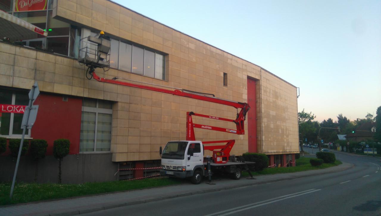 Pracownik w żółtym kasku na podnośniku koszowym przy elewacji budynku z płytek, widoczna ciężarówka z podnośnikiem Oile&Steel na tle ulicy.
