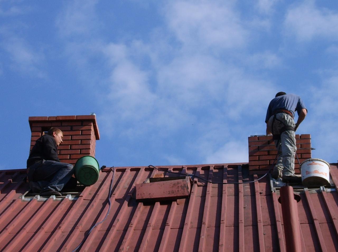 Dwóch dekarzy na czerwonym dachu, pracujących przy murowanych kominach na tle błękitnego nieba. Jeden siedzi, drugi stoi przy kominie.