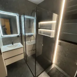 Nowoczesna łazienka z kabiną prysznicową typu walk-in, umywalką z szafką i lustrem z podświetleniem LED. Ściany i podłoga wykończone w ciemnym, nakrapianym kamieniu.