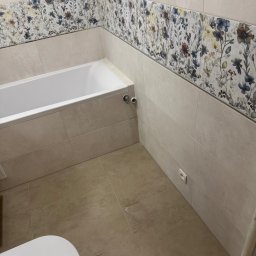 Akazius Professional - Wykończona łazienka z beżowymi płytkami i dekoracyjnym pasem kwiatowym. Widoczna wanna, fragment toalety i gniazdko elektryczne. Neutralne kolory i nowoczesny design.