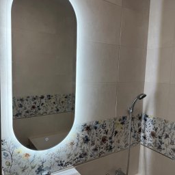 Akazius Professional - Wykończona łazienka w Białymstoku: owalne lustro LED, umywalka i bateria, płytki z motywem kwiatowym, chromowana armatura natryskowa.