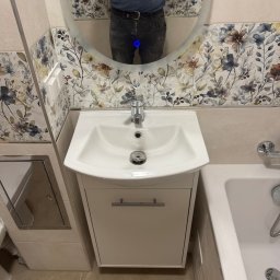 Akazius Professional - Nowoczesna łazienka po remoncie: biała umywalka z szafką, bateria, owalne lustro z widocznym odbiciem osoby, płytki z motywem kwiatowym i beżowe płytki podłogowe.