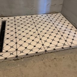 Akazius Professional - Wykończona posadzka kabiny prysznicowej z odpływem liniowym. Białe płytki z czarnym wzorem geometrycznym, szare, surowe ściany i podłoga w tle.