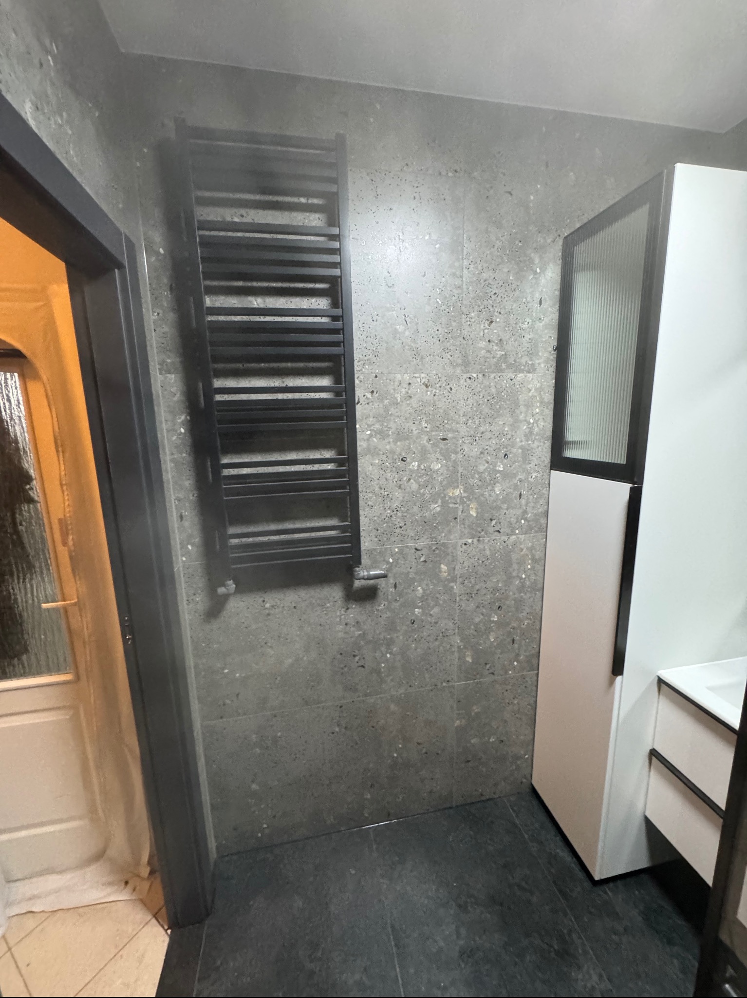 Wykończona łazienka w szarościach: ciemna podłoga, ściany z efektem lastryko, czarny grzejnik drabinkowy i biała szafka z umywalką.