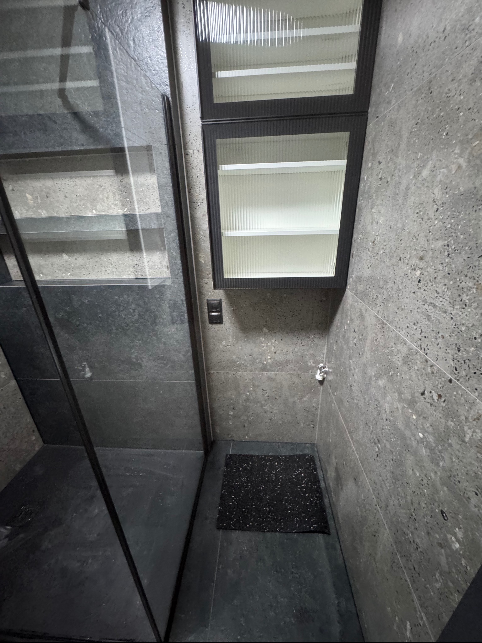 Nowoczesna kabina prysznicowa z szarymi płytkami imitującymi kamień, czarnym brodzikiem i szafką z ryflowanym szkłem. Minimalistyczny design.