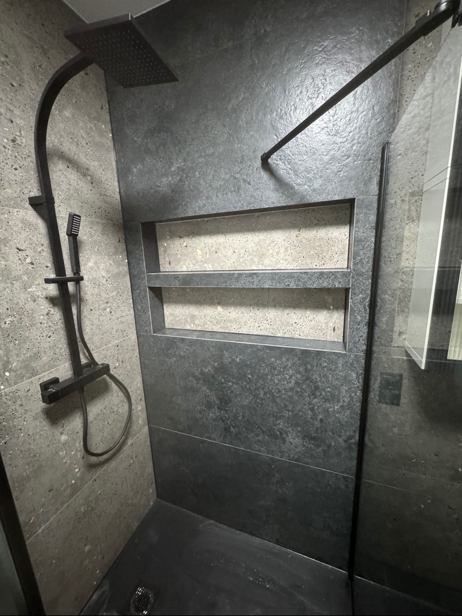 Nowoczesna kabina prysznicowa z czarną armaturą i szarymi płytkami imitującymi kamień, z wnękami na kosmetyki. Minimalistyczny design.