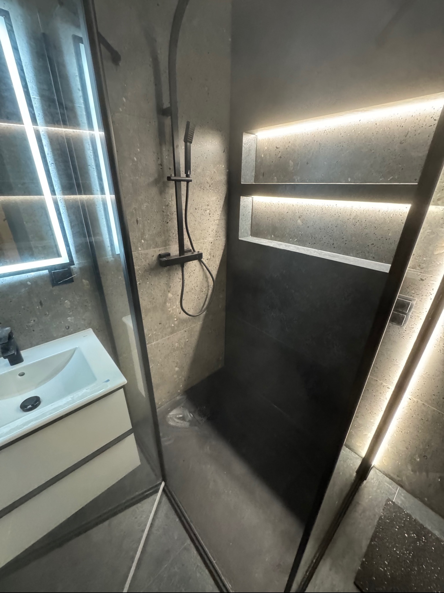 Nowoczesna kabina prysznicowa walk-in z czarną armaturą i podświetlanymi wnękami, obok umywalka z szafką. Aranżacja łazienki w szarościach z akcentami led.