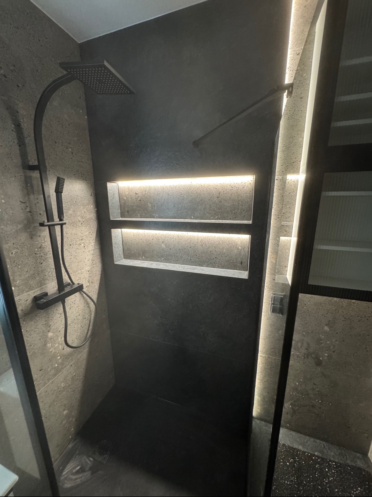 Nowoczesna kabina prysznicowa z czarną armaturą i wnękami podświetlonymi LED, wykończona szarą, kamienną płytką. Minimalistyczny design, ciemna kolorystyka, eleganckie oświetlenie.