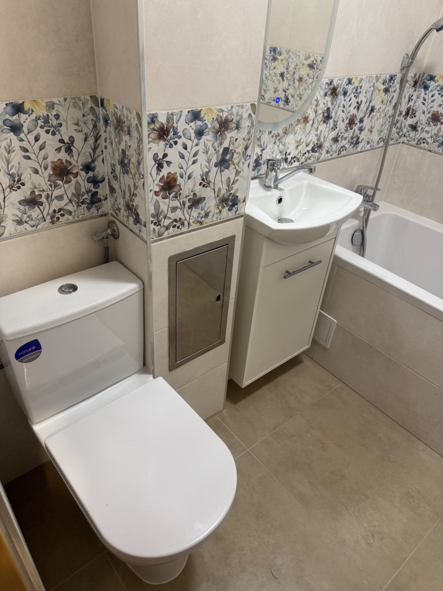 Biała toaleta Cersanit i umywalka z szafką w wykończonej łazience. Na ścianach płytki z motywem kwiatowym, beżowa podłoga. Widoczna wanna i lustro.