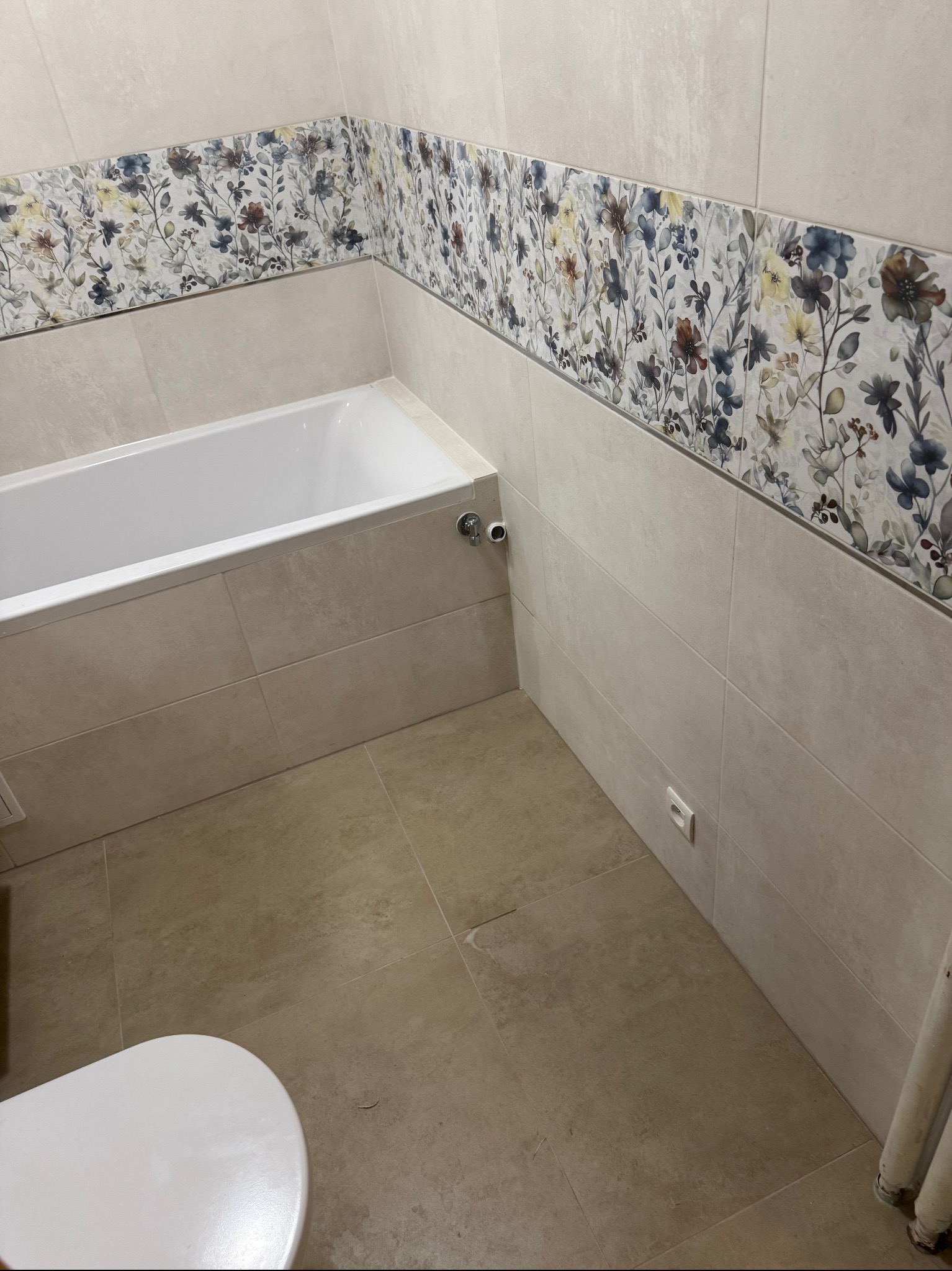 Wykończona łazienka z beżowymi płytkami i dekoracyjnym pasem kwiatowym. Widoczna wanna, fragment toalety i gniazdko elektryczne. Neutralne kolory i nowoczesny design.