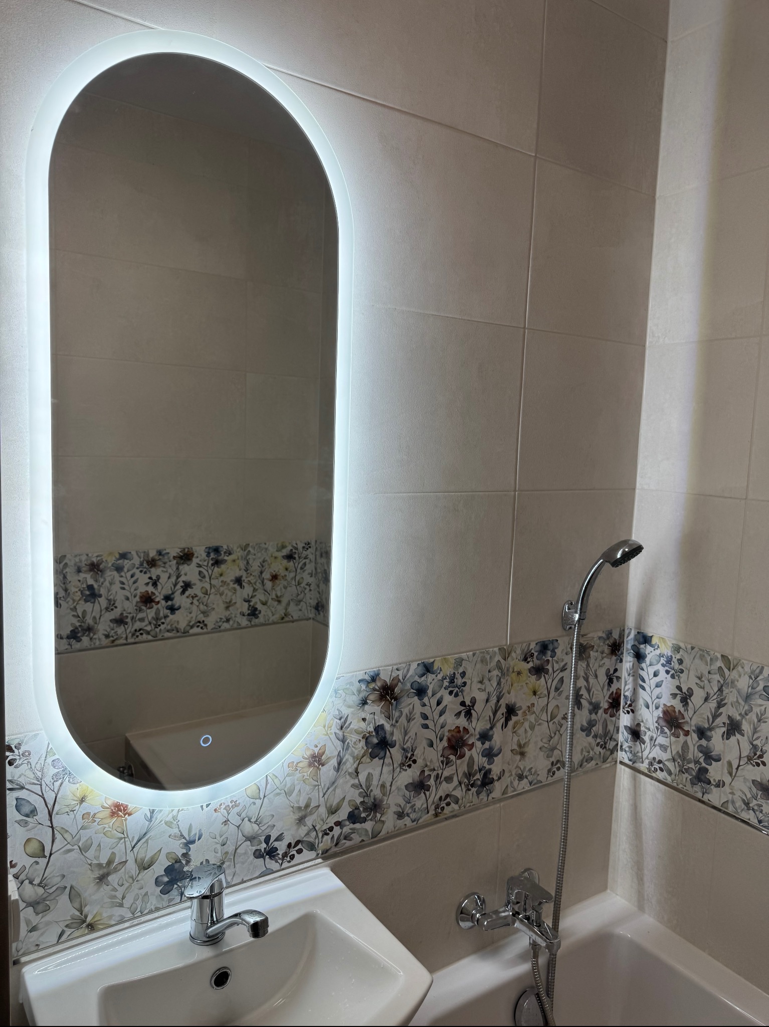 Wykończona łazienka w Białymstoku: owalne lustro LED, umywalka i bateria, płytki z motywem kwiatowym, chromowana armatura natryskowa.