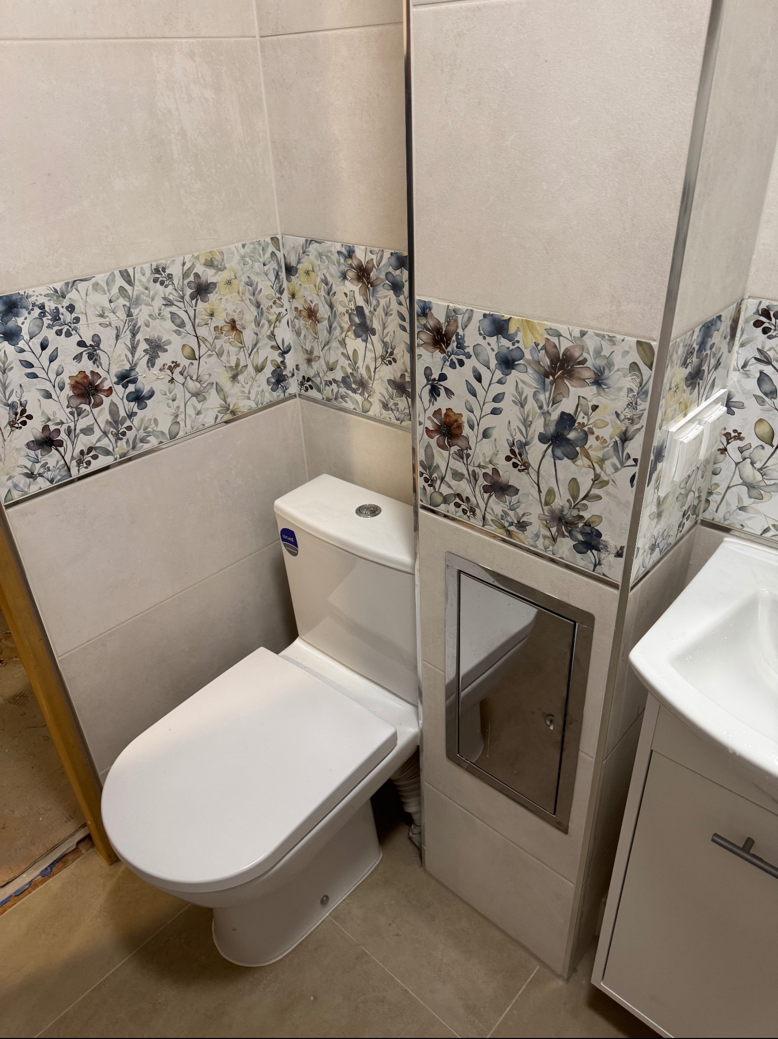 Wykończona łazienka: biała toaleta i umywalka z szafką, kremowe płytki z kwiatowym pasem dekoracyjnym. Widoczny fragment podłogi przed położeniem płytek.