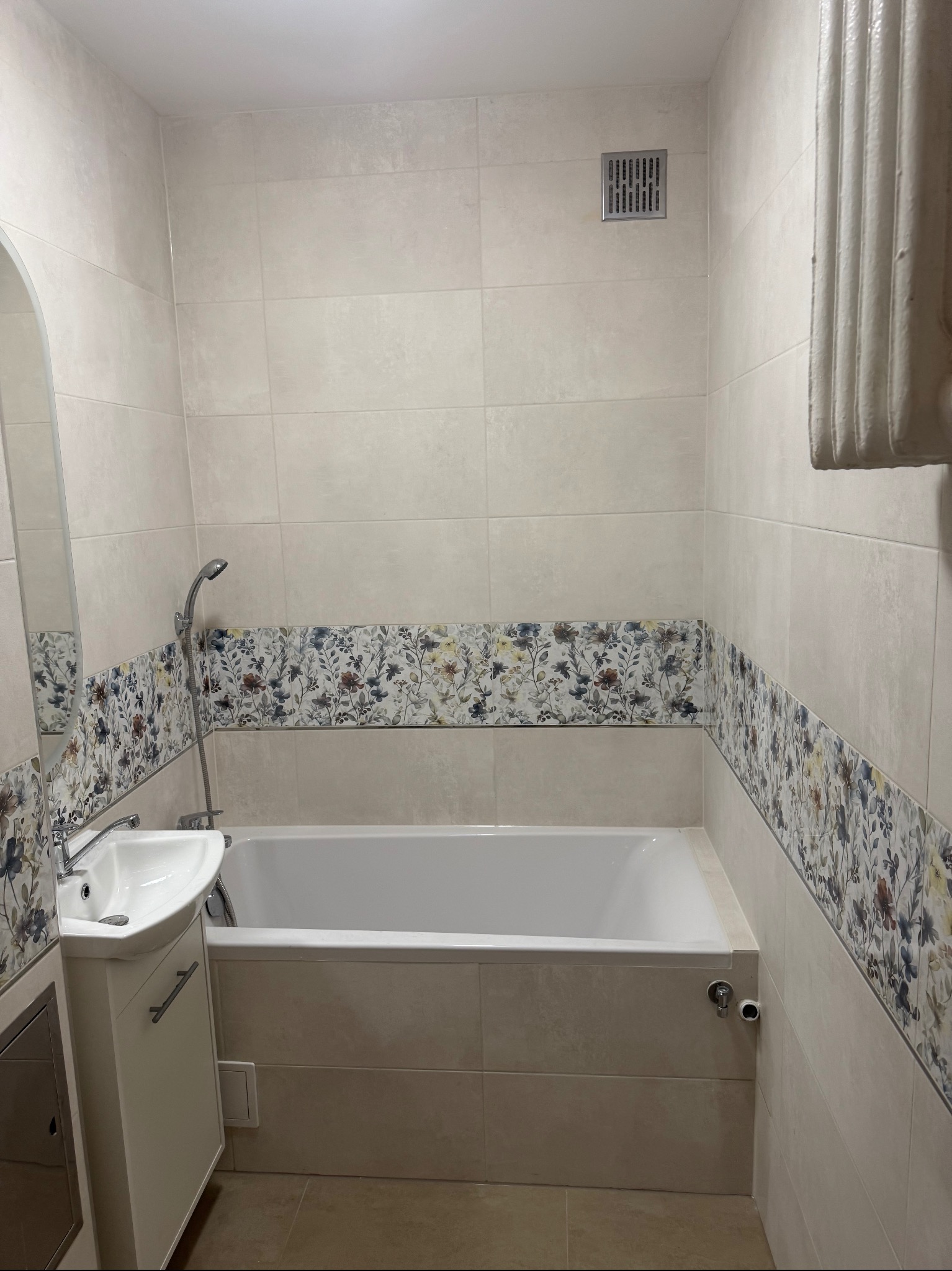 Jasna łazienka po remoncie: beżowe płytki, dekoracyjny pas z motywem kwiatowym, biała wanna i umywalka z szafką. Grzejnik żeberkowy po prawej.