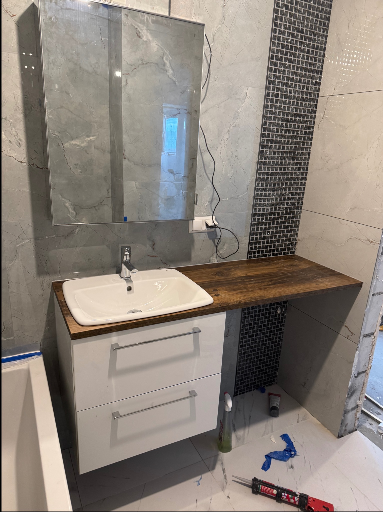 Nowoczesna łazienka z umywalką nablatową na białej szafce z szufladami, drewnianym blatem i lustrem. Ściany wyłożone płytkami imitującymi marmur i mozaiką.