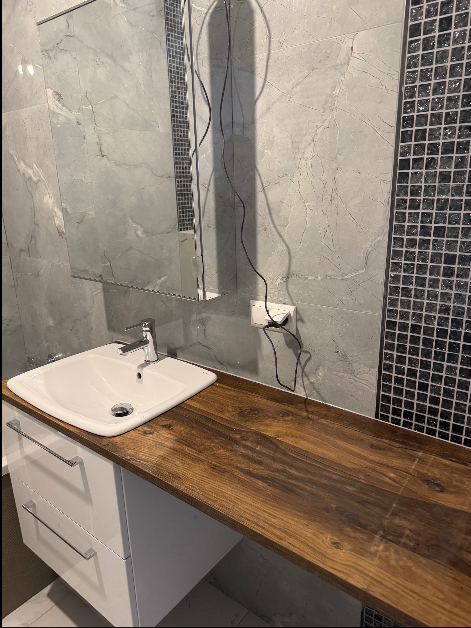 Wykończona łazienka: biała umywalka na drewnianym blacie, lustro z szafką, ściany z szarych płytek imitujących marmur i dekoracyjna mozaika z czarnych kwadratów.