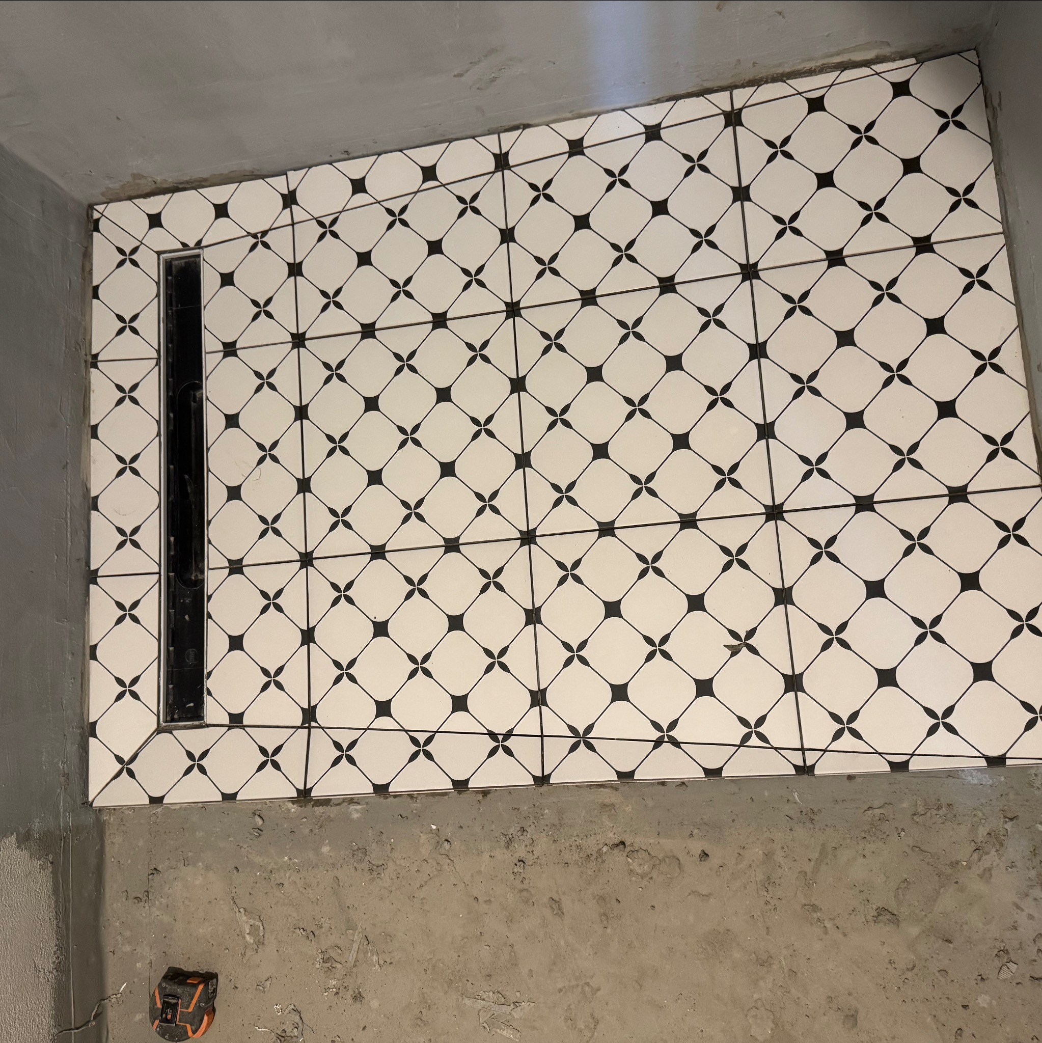 Ułożone białe płytki podłogowe z czarnym wzorem i odpływem liniowym w kabinie prysznicowej. Widoczny surowy beton posadzki i fragment poziomicy laserowej.