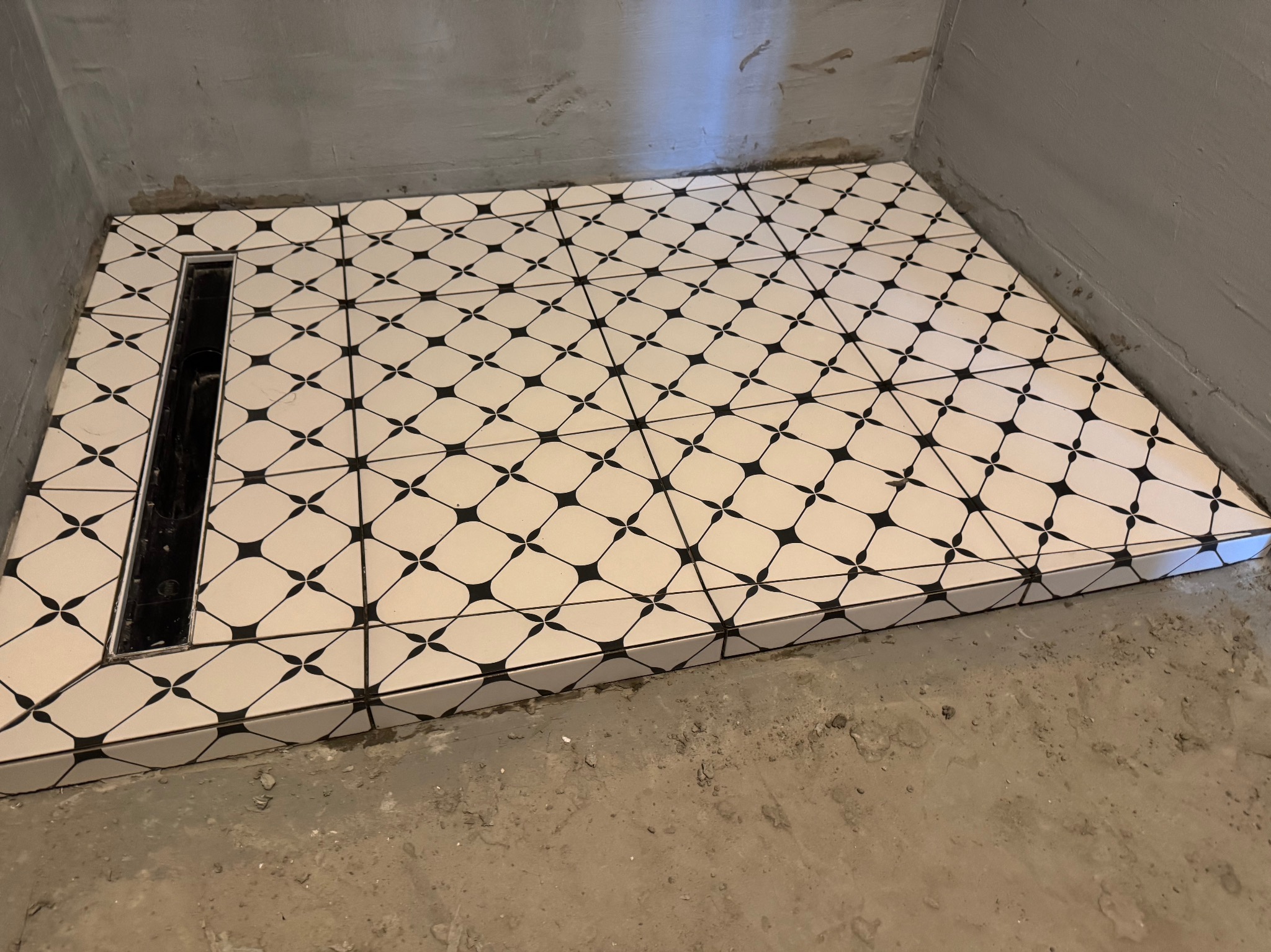 Wykończona posadzka kabiny prysznicowej z odpływem liniowym. Białe płytki z czarnym wzorem geometrycznym, szare, surowe ściany i podłoga w tle.