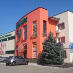 Dwupiętrowy budynek z pomarańczową fasadą i zielonym napisem, widoczne zaparkowane samochody i znak parkingu.