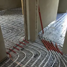 Montaż rur ogrzewania podłogowego na folii izolacyjnej z siatką, widoczny poziomica oparta o surową ścianę.