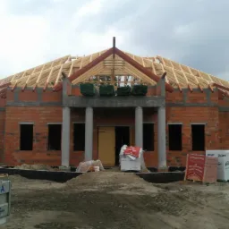 Budowa domu jednorodzinnego w stanie surowym otwartym: ceglana konstrukcja, drewniana więźba dachowa, kolumny wejściowe i materiały budowlane na placu.