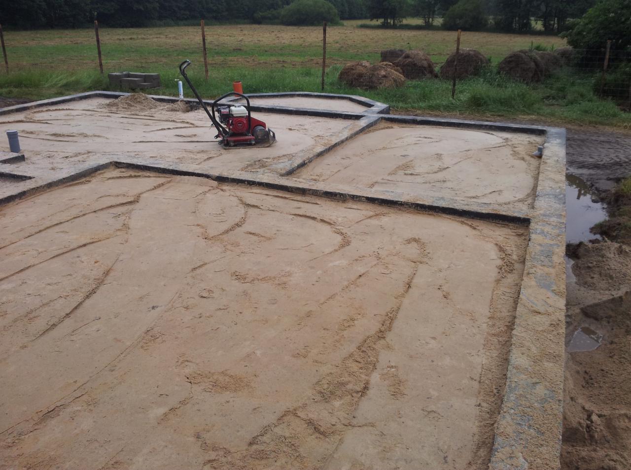 Etap przygotowania podłoża pod wylewkę z użyciem zagęszczarki, widoczne czarne obramowanie i piasek. W tle teren zielony i hałdy siana.