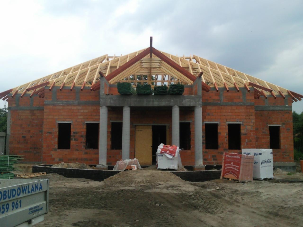 Budowa domu jednorodzinnego w stanie surowym otwartym: ceglana konstrukcja, drewniana więźba dachowa, kolumny wejściowe i materiały budowlane na placu.