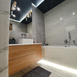 Usługi budowlane Makowski - Nowoczesna łazienka z marmurowymi płytkami, drewnianą szafką pod umywalką, czarnym sufitem z oświetleniem LED i szarym dywanikiem podłogowym.