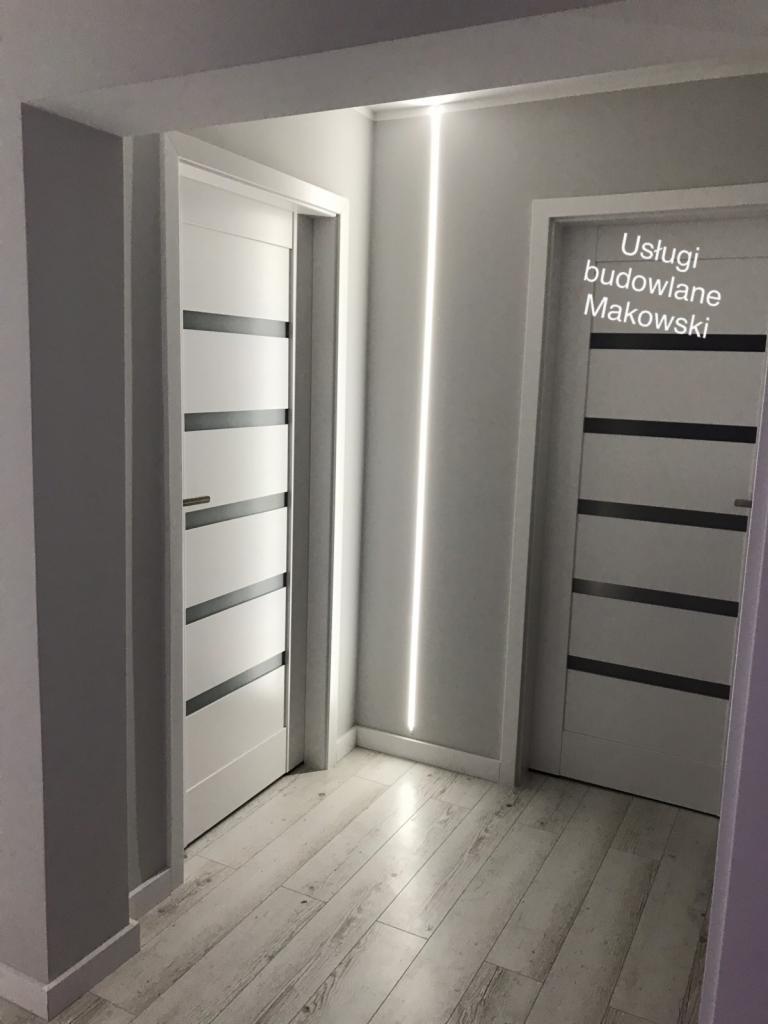 Dwa białe, nowoczesne skrzydła drzwiowe z poziomymi, szarymi wstawkami, zamontowane w szarym korytarzu z podłogą imitującą jasne drewno, oświetlone pionową listwą LED.