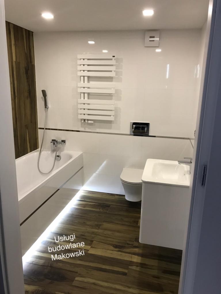 Nowoczesna łazienka z drewnopodobną podłogą, białą armaturą i podświetleniem LED pod wanną, widok zza uchylonych drzwi.