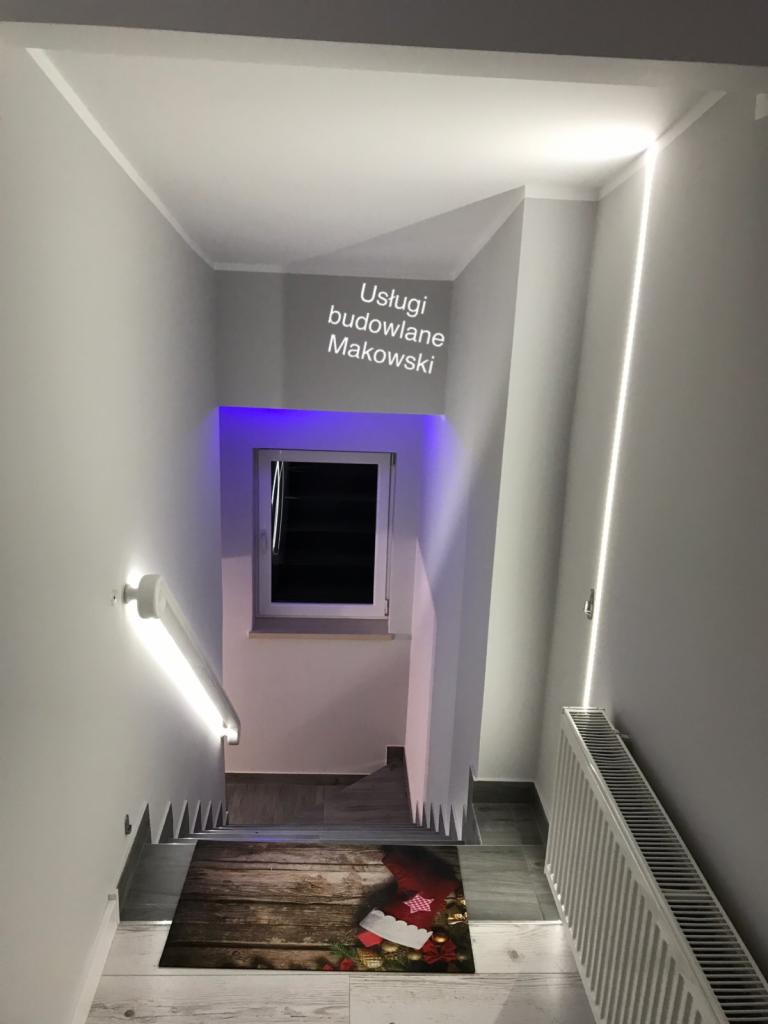 Klatka schodowa z oświetleniem LED, widok z góry na dywanik świąteczny na schodach, grzejnik na ścianie po prawej stronie, okno z fioletowym podświetleniem w tle.