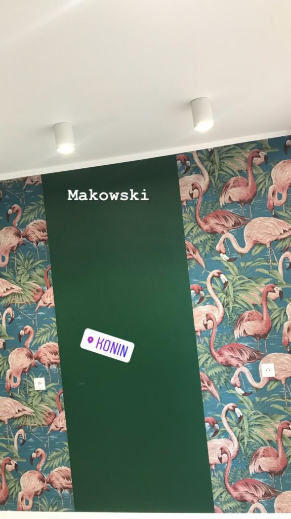 Ściana z tapetą w flamingi przylegająca do ciemnozielonej, pionowej powierzchni z napisem 'Makowski' i naklejką lokalizacyjną 'Konin', oświetlona dwoma białymi reflektorami sufitowymi.
